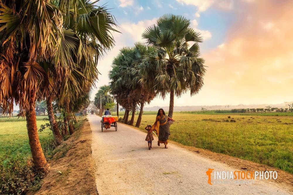 Rural woman with her child walking along an unpaved village road at sunset